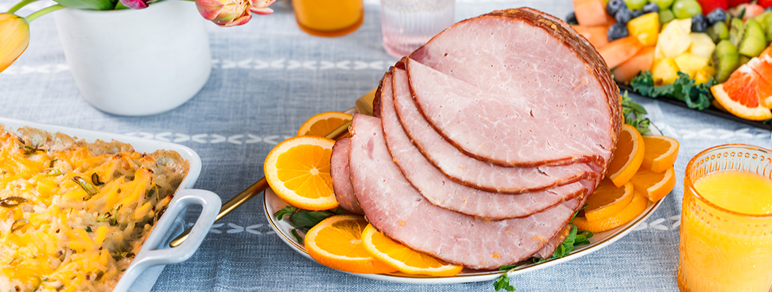 a sliced ham on a platter set on a table with a casserole, orange juice and sliced fruit platter.