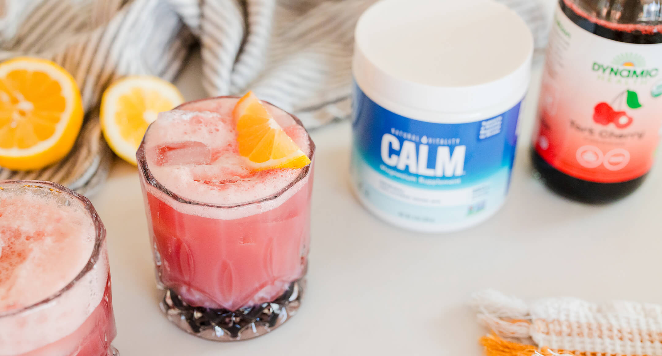 A glass with a drink and orange slice in it sits on a table top next to a container of CALM magnesium supplement and tart cherry juice.