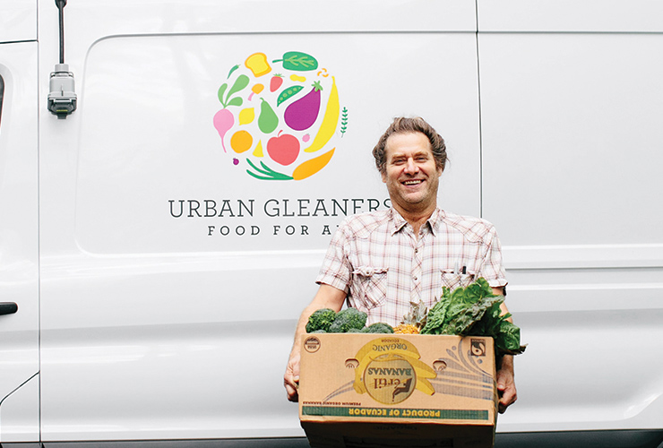 Smiling person holding a box of fresh vegetables in front of a white van with the Urban Gleaners logo.