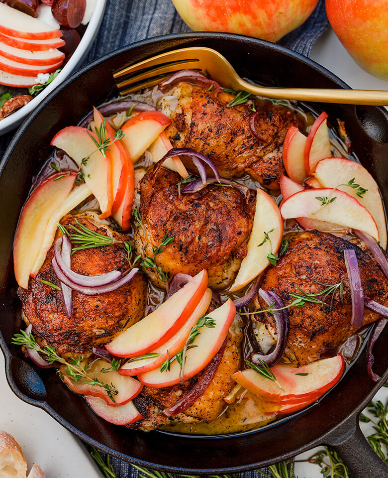 A cast-iron pan with roasted chicken thighs, sliced apples, and red onion.