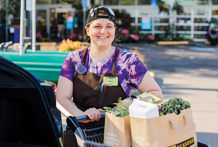 a smiling staff member wearing a backwards baseball hat and pushing full bags of groceries in a shopping cart in the store parking lot.