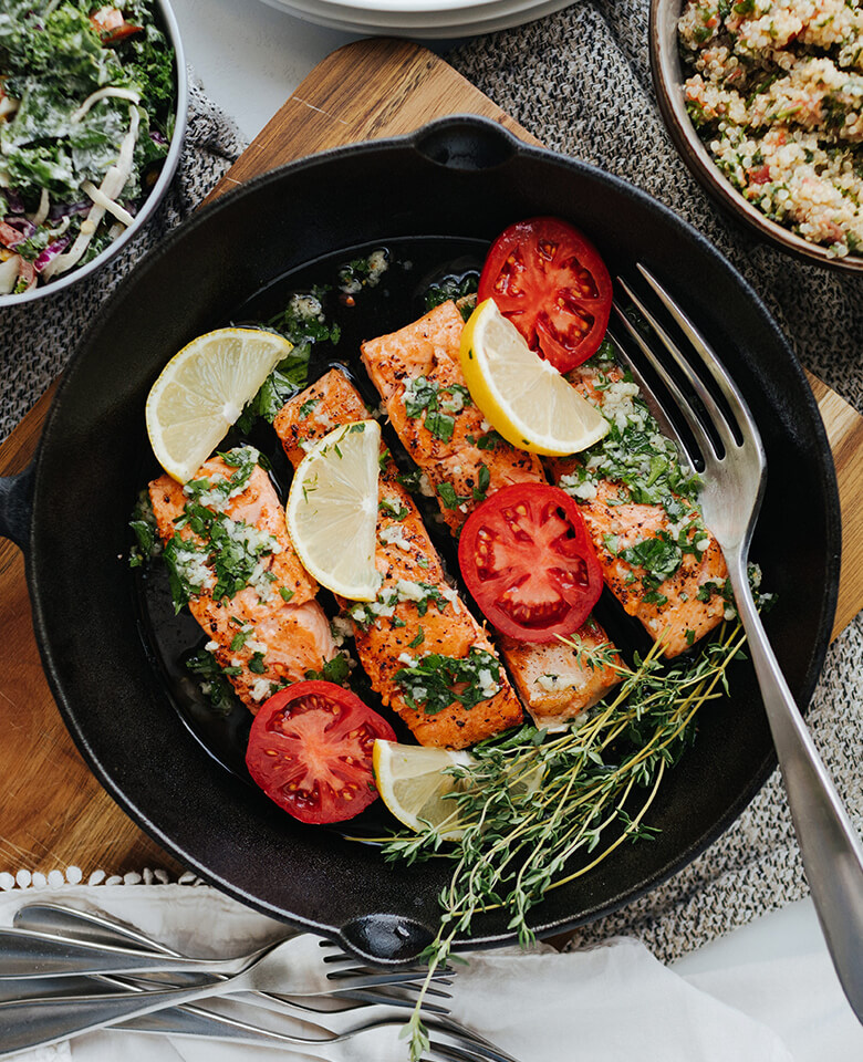 Salmon fillets in a cast-iron pan with lemon, tomato, and fresh herbs.