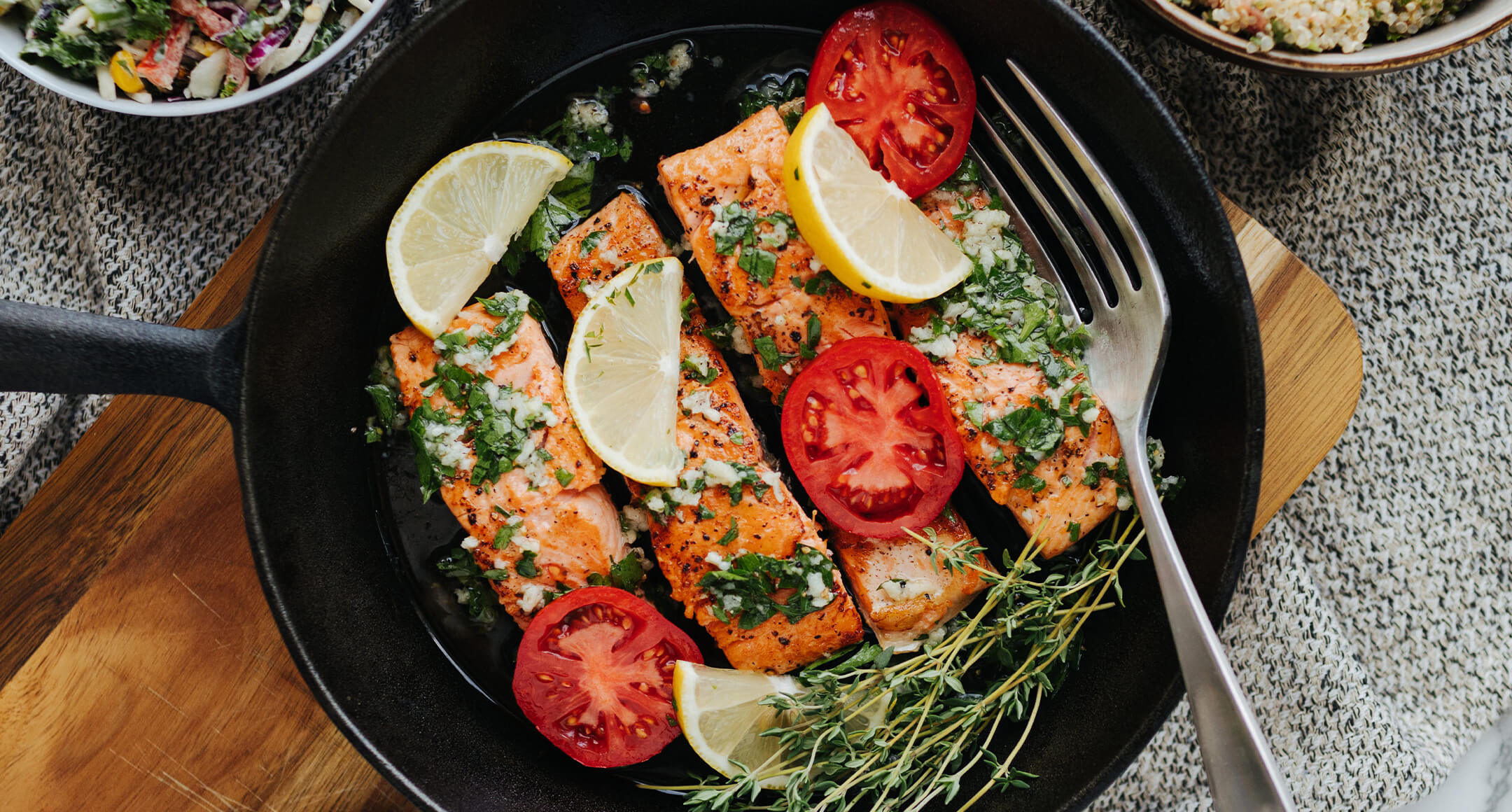 Salmon fillets in a cast-iron pan with sliced tomato and fresh herbs.