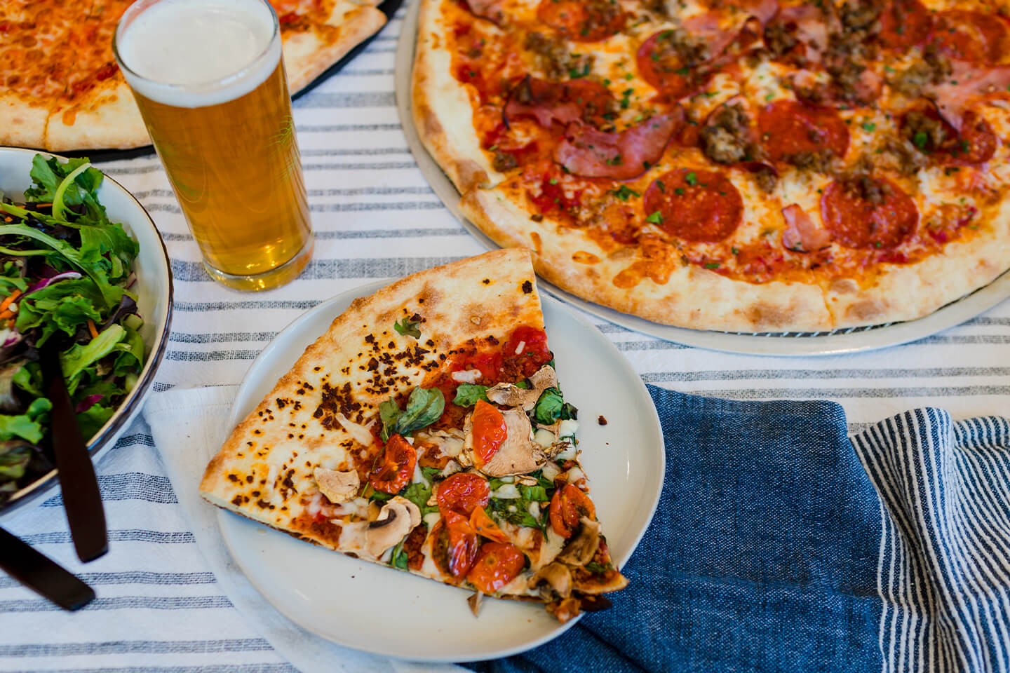 Picnic setting featuring a whole pizza, side salad, beer, and slice of pizza on a plate.