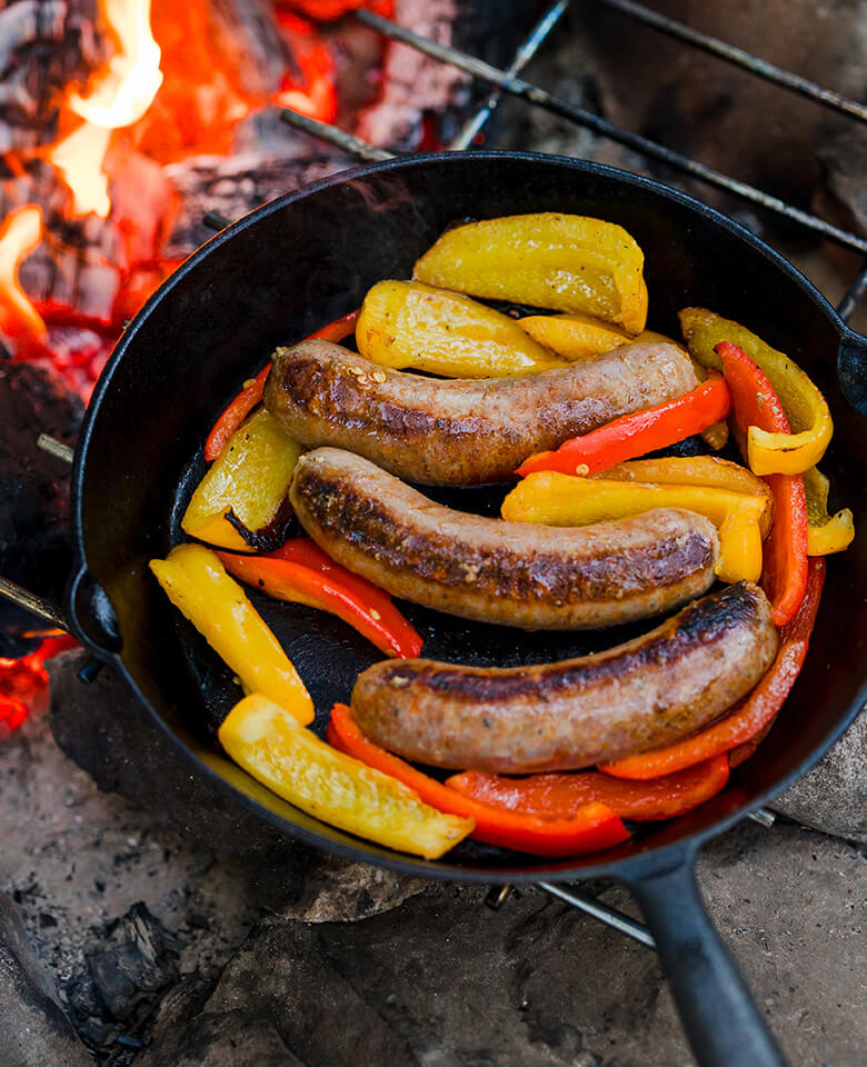 Sausage links and peppers in a cast iron skillet grilling over a camp fire
