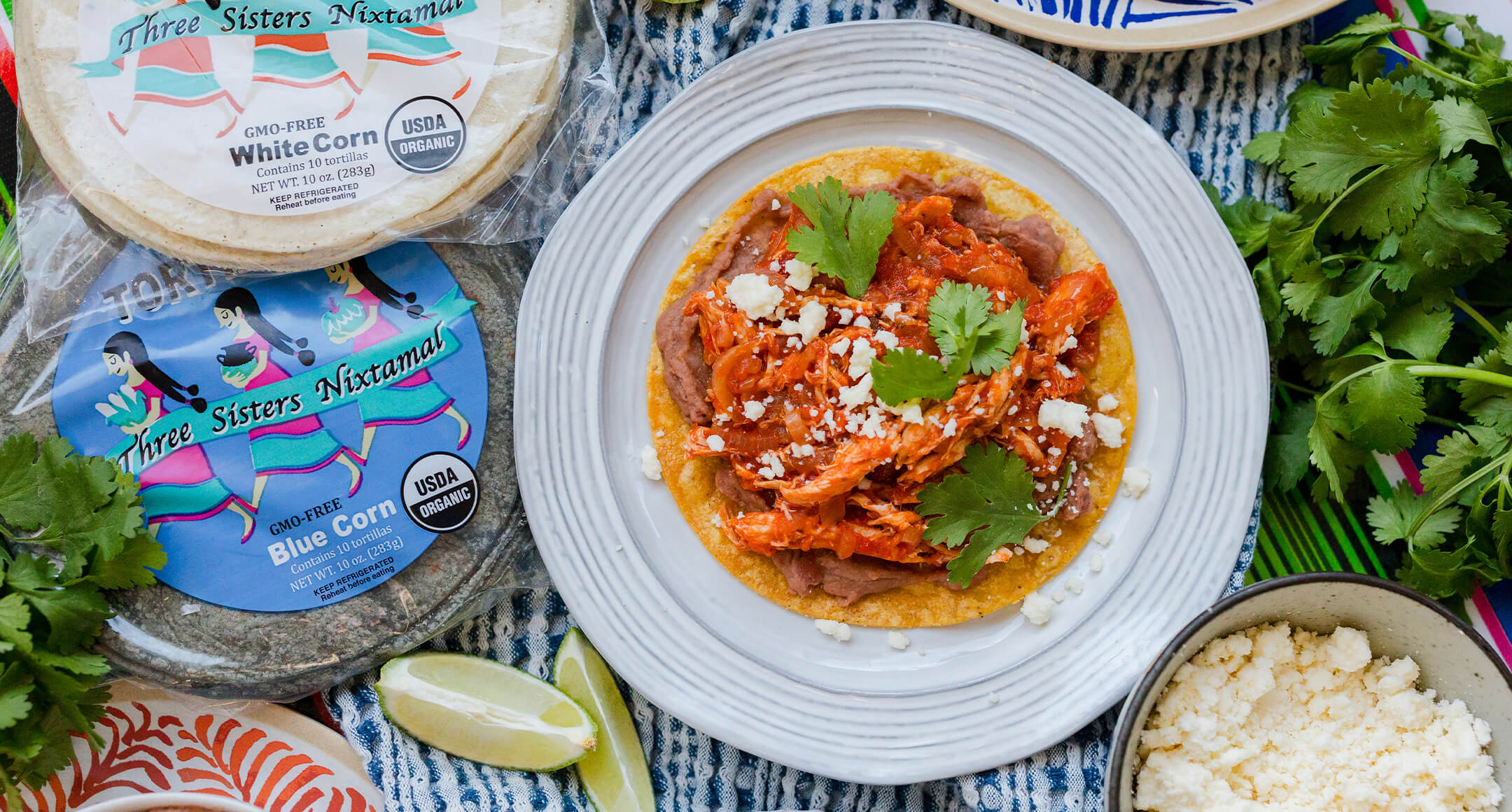 A chicken Tinga tostada on a plate next to packages of Three Sisters Nixtamal corn tortillas.