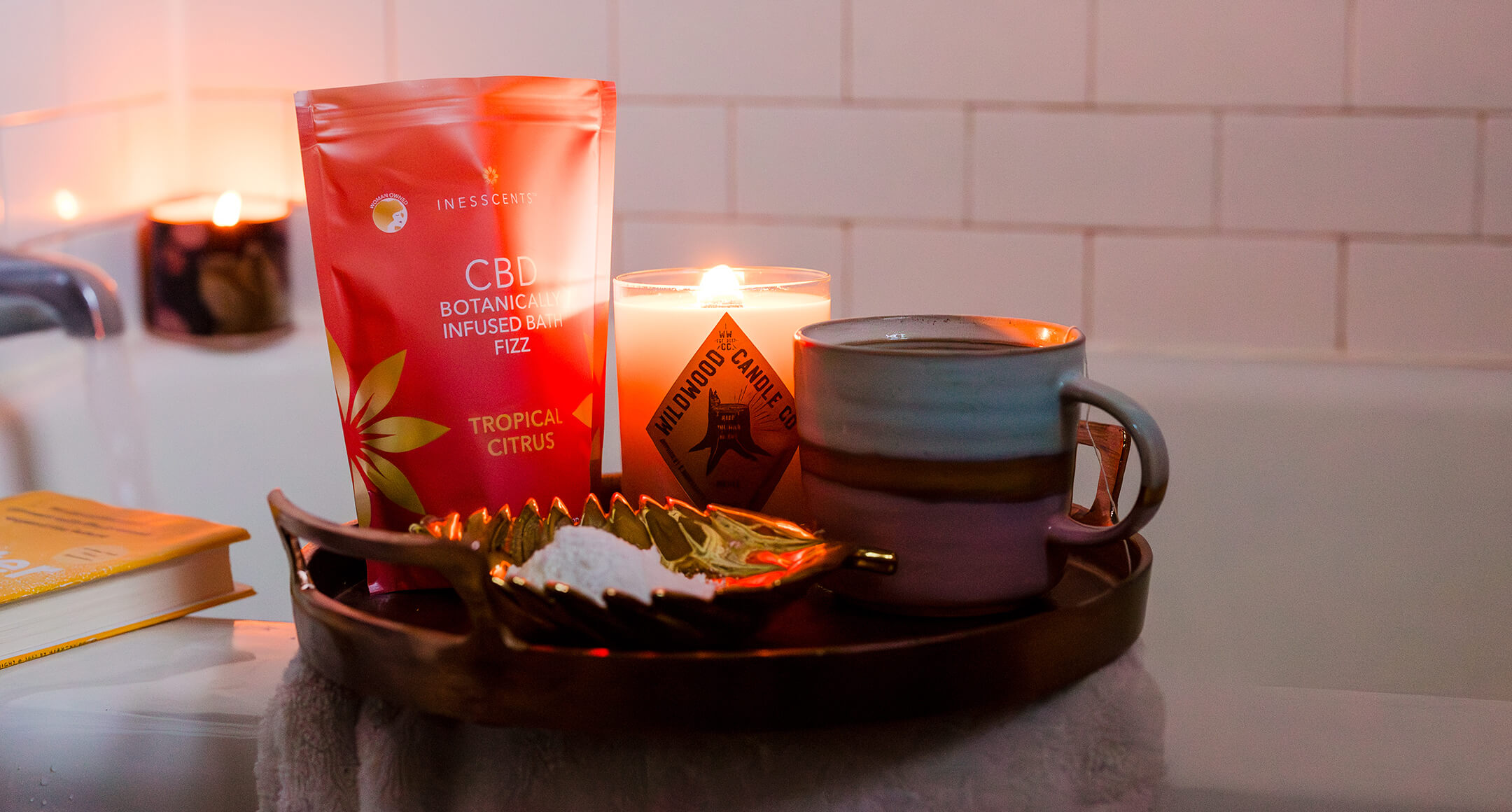 A tray with CBD bath salts, a mug of tea and a lit candle with a bath tub and book in the background.