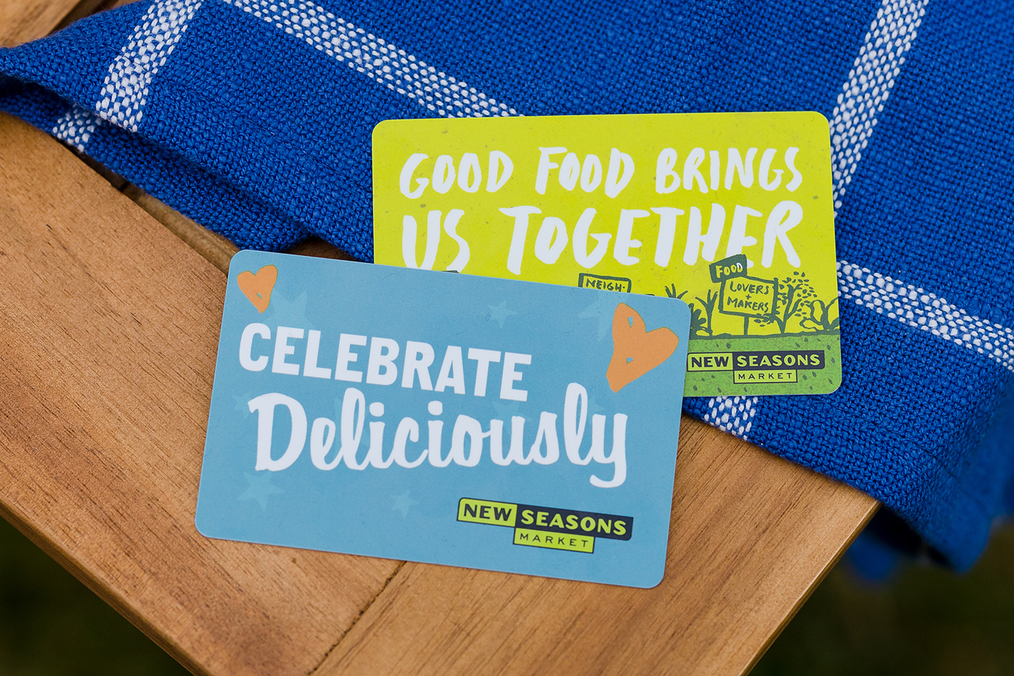 New Seasons Market gift cards with the words "good food brings us together" and "celebrate deliciously" placed on a table with a blue towel underneath.