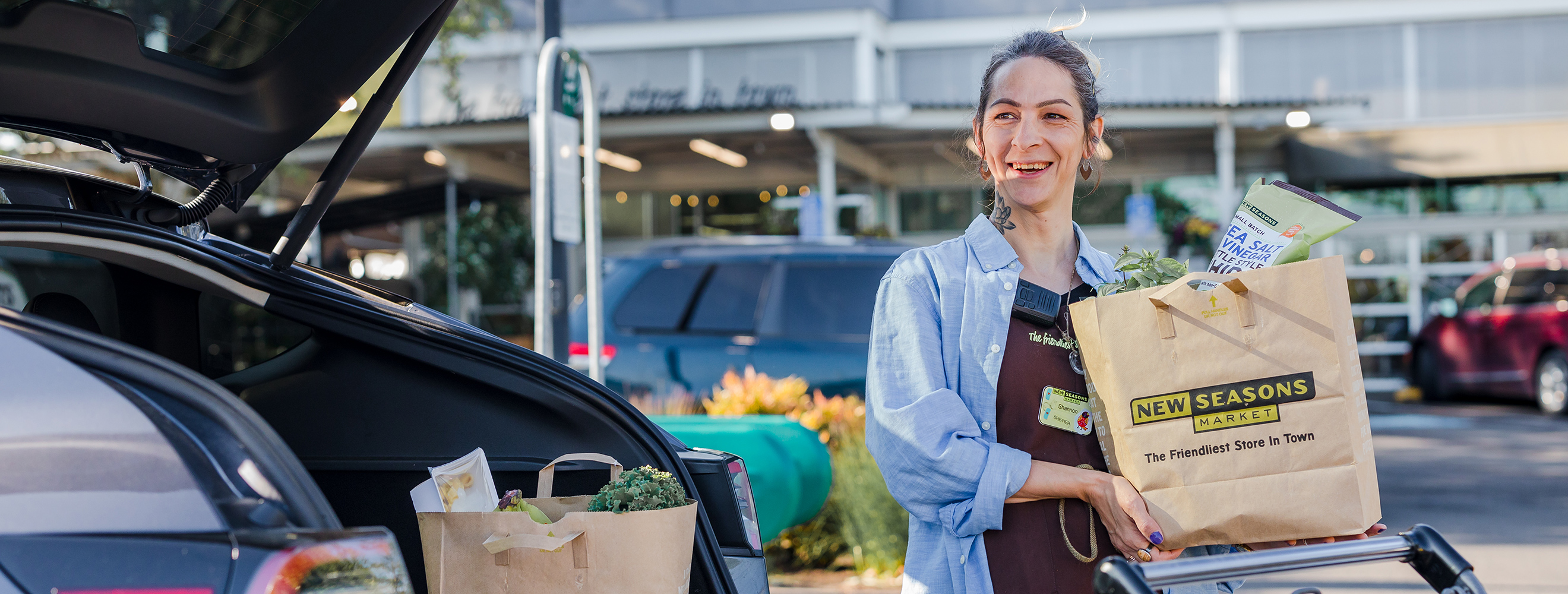 a smiling New Seasons Market staff member carrying a bag of groceries to the trunk of a car.