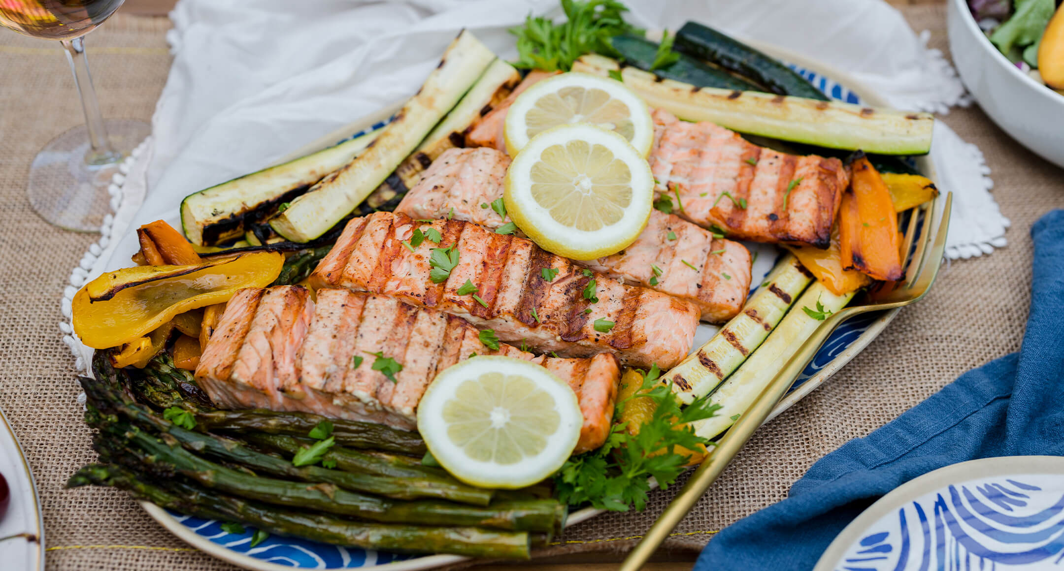 Grilled salmon fillets on a plate topped with sliced lemon and surrounded by grilled zucchini and asparagus spears. 