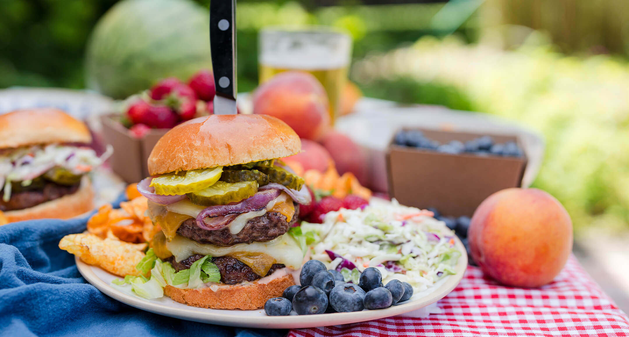 A fully composed cheeseburger on a plate with sides of chips, coleslaw, and fresh fruit. 