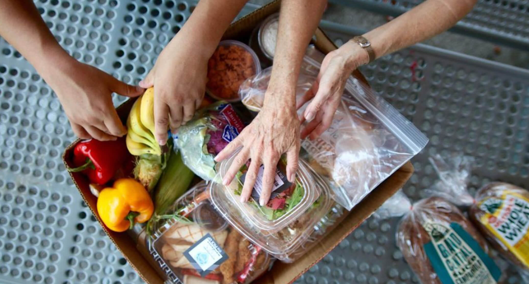 A close-in bird’s-eye image showing four hands loading food into a cardboard box.