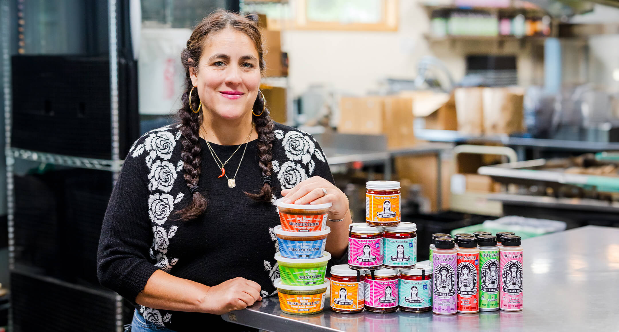 Image of Nikki Guerrero, owner of Hot Mama in Portland, OR, standing next to her products: salsa, sauces, and chili oils.