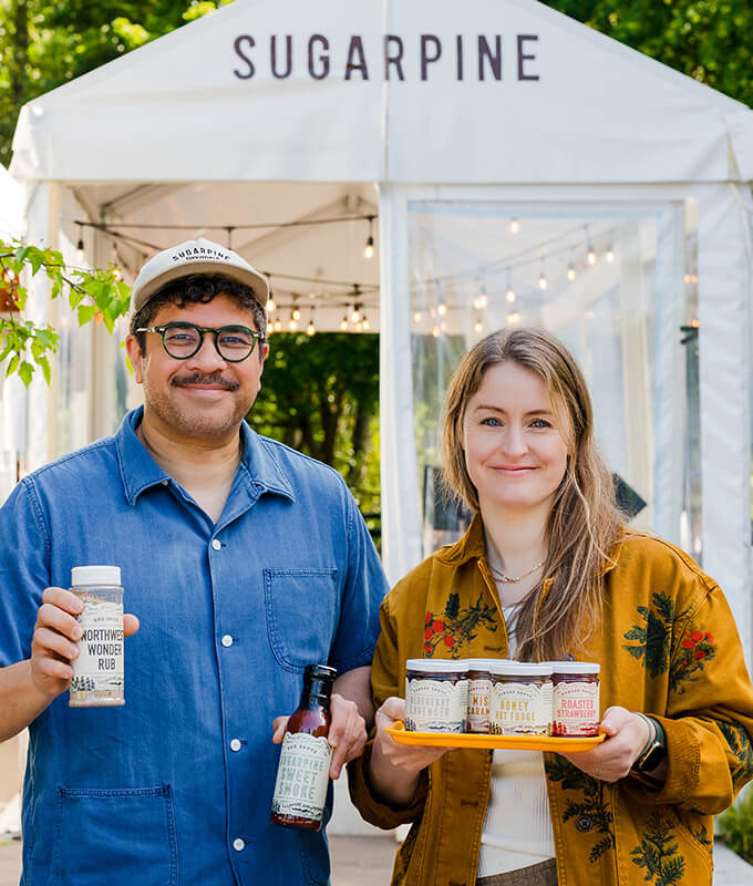two people standing in from of a tent with the words "sugarpine" and holding bottles of their BBQ sauce products.